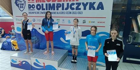 Powiększ grafikę: dziewczynki na podium po zawodach pływackich, w tle baner zawodów
