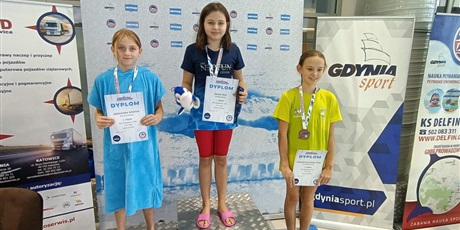Powiększ grafikę: trzy uczennice - pływaczki na podium z dyplomami i medalami na szyjach na tle reklamowych banerów