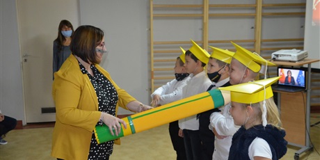 Powiększ grafikę: pani wicedyrektor dotyka ramienia ucznia dużym, żółtym ołówkiem